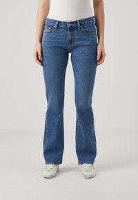 Jeans a zampa d'elefante in denim blu con vita alta, tasche frontali e dettagli di cucitura sottili. Indossati con sneakers bianche.