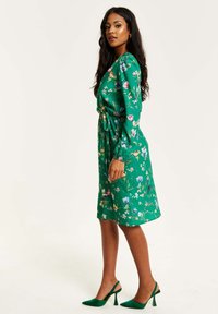 Green floral print dress with long sleeves, cinched waist, and knee-length hem. Complemented by green high-heeled shoes.