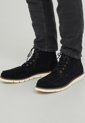 Botas de tobillo de ante navy con suelas de goma blanca en contraste, seis ojales para los cordones y un diseño de punta plana. Acabado texturizado con costuras visibles.
