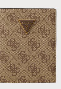 Brown patterned canvas wallet featuring gold triangular Guess logo, distinctive monogram designs, and contrasting dark border stitching.