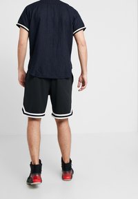 Sorte atletikshorts med hvid kant og struktureret stof, kombineret med en marineblå kortærmet skjorte med hvide striber. Sorte sneakers med røde detaljer.