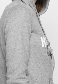 Grå zip-hoodie i mjukt material, med huva, dragsko och vit tryck på bröstet, i avslappnad passform.