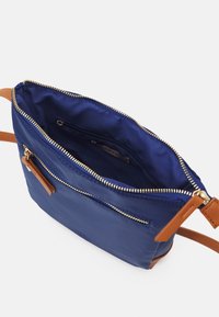Námořnická modrá nylonová kabelka s hnědými koženými detaily, horním zipovým uzávěrem a vnitřní kapsou. Hladká textura, obdélníkový tvar a nastavitelné ramenní popruhy.