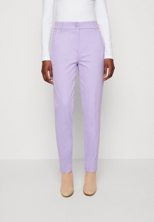 Personne portant un pantalon slim lavande, un haut blanc à manches longues et des bottines beige, debout devant un fond uni.