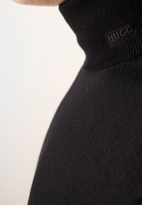 Schwarzer Stehkragenpullover aus Strick, mit einem kleinen geprägten Logoetikett am Kragen. Glatte Textur mit tailliertem Schnitt.