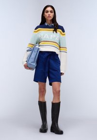 Maglione a maglia con strisce blu, gialle e verdi, con la scritta "NAPA". Abbinato a pantaloni corti blu navy e stivali in gomma neri al ginocchio.