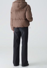 Brązowa kurtka puchowa z kapturem, z teksturowanym kołnierzem i elastycznymi mankietami, zestawiona z ciemnymi, prostymi spodniami dżinsowymi.