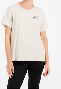 Jasno beżowa bawełniana koszulka z okrągłym dekoltem i krótkimi, zrolowanymi rękawami. Z przodu, po lewej stronie, znajduje się kolorowe logo "PROTEST".