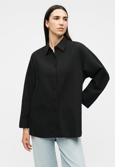 Γυναίκα με ίσια σκούρα μαλλιά, που φοράει μια φαρδιά μαύρη πουκάμισα με κουμπιά και ανοιχτό μπλε τζιν, ποζάροντας με το ένα χέρι στη μέση της.