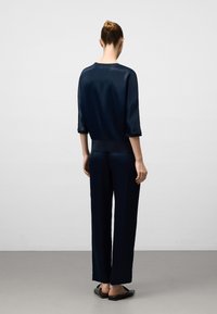 Kvinna står med ryggen mot oss, iförd en marinblå satinblus med trekvartslånga ärmar, matchande vida byxor och svarta slip-on skor.