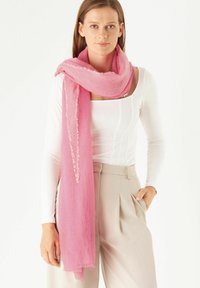 Rosa Schal aus leichtem Stoff mit fransigem Rand. Er umschließt den Hals und ergänzt ein weißes Oberteil und beigefarbene Hosen.