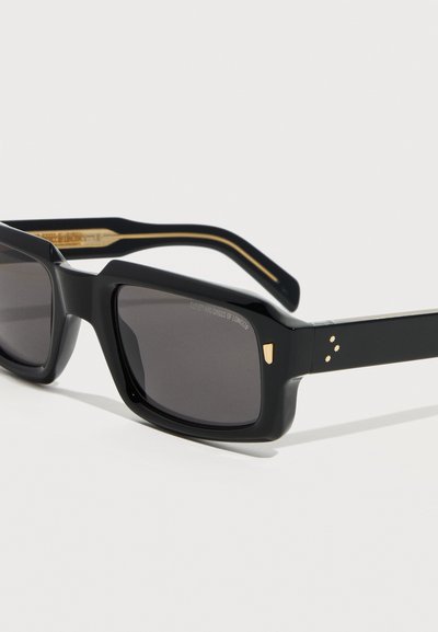 Μαύρα ορθογώνια γυαλιά ηλίου με σκουρόχρωμους φακούς, χρυσές λεπτομέρειες στις γωνίες και στα μπράτσα, γωνιακή κοντινή λήψη σε λευκό φόντο.