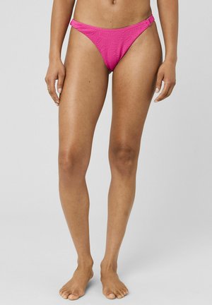 Femme portant un bas de bikini texturé rose vif, debout pieds nus sur un fond gris uni.