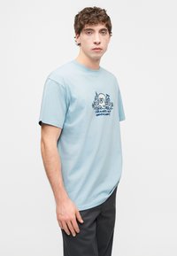 Ανοιχτό μπλε βαμβακερό μπλουζάκι με κοντά μανίκια και στρογγυλή λαιμόκοψη, με σχέδιο σε ναυτικό μπλε απόχρωση που απεικονίζει ένα χταπόδι και κείμενο στο μπροστινό μέρος.