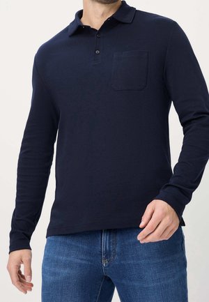 Mann trägt ein marineblaues langärmeliges Poloshirt mit einer Brusttasche und blaue Jeans aus Denim vor einem schlichten Hintergrund.