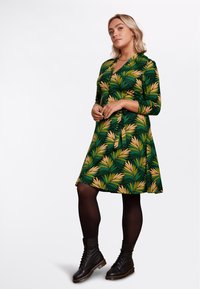 Abito verde con stampa a foglia in giallo e verde, design avvolgente, maniche tre quarti, abbinato a stivali neri alla caviglia e collant.