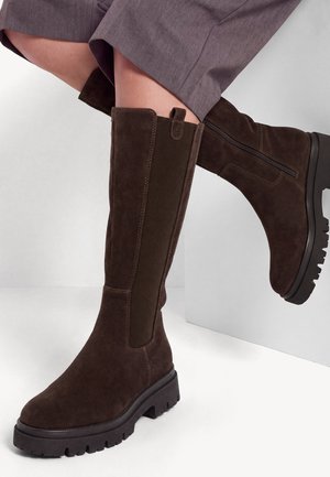 Bottes en daim marron montantes avec fermeture éclair et panneau latéral extensible, portées avec un pantalon gris, montrant les détails de la semelle et du talon.