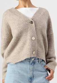 Beżowy sweter na drutach z dekoltem w kształcie litery V, z złotymi guzikami i prążkowanymi mankietami, zestawiony z jasnoniebieskim deninem. Miękka faktura, luźny krój.