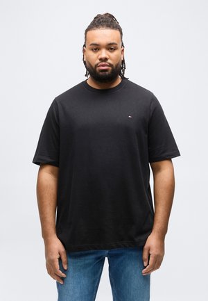 Homme aux cheveux tressés et à la barbe, portant un t-shirt noir à manches courtes et un jean bleu, debout devant un fond gris clair uni.