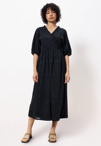 Schwarzes Wickelkleid mit strukturiertem Blumenmuster, V-Ausschnitt, Puffärmeln und tailliertem Schnitt, das bis zur Mitte der Waden reicht. Kombiniert mit olivfarbenen Sandalen.