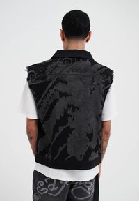 Gilet di denim nero con motivo floreale in rilievo, orlo sfrangiato e colletto, indossato su una t-shirt bianca. Presenta dettagli grigi a contrasto.