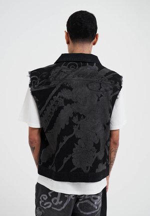 Gilet di denim nero con motivo floreale in rilievo, orlo sfrangiato e colletto, indossato su una t-shirt bianca. Presenta dettagli grigi a contrasto.