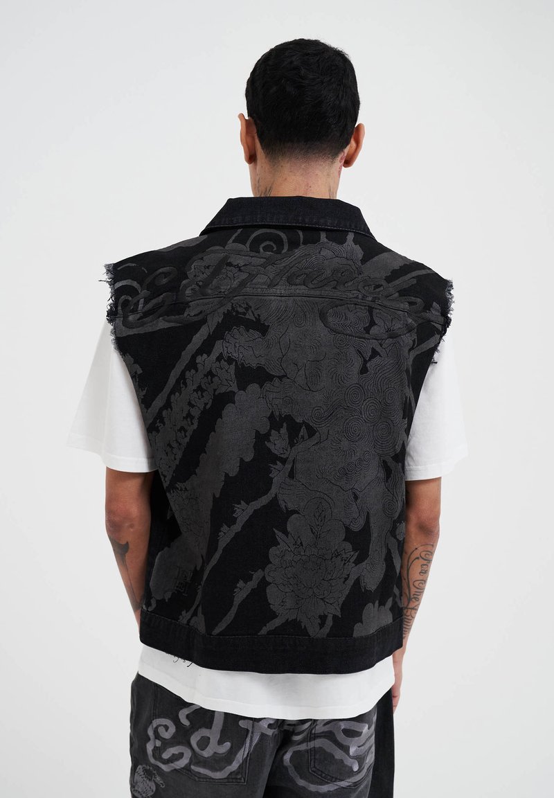 Gilet di denim nero con motivo floreale in rilievo, orlo sfrangiato e colletto, indossato su una t-shirt bianca. Presenta dettagli grigi a contrasto.