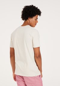 Lys beige kortærmet t-shirt med rund halsudskæring, glat tekstur og subtilt mærke i kanten. Bæret med lyserøde fløjlsshorts.