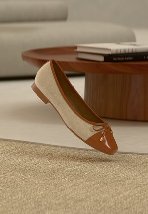 Beige og brun balletflat med sløjfedetalje, placeret nær et rundt træ-sofabord med stabler af bøger på et beige tæppe.