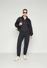 Pullover in pile nero con zip a quarti, abbinato a pantaloni jogger coordinati. Il modello indossa scarpe da ginnastica beige e occhiali da sole. Inclusa una borsa da viaggio nera.