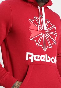 Sudadera roja de tejido suave, con un prominente logo blanco de Reebok y un diseño geométrico de estrella en el frente. Incluye capucha con cordón.