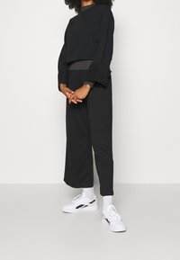 Top corto negro con mangas anchas superpuesto a una camiseta de manga larga gris ajustada, combinado con pantalones negros holgados y zapatillas blancas.