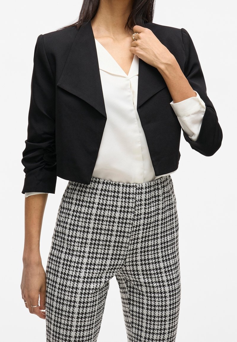Zwarte cropped blazer met een reverskraag, gemaakt van gladde stof. Gecombineerd met een witte blouse en zwart-wit geruite pantalons met een pied-de-poule patroon.