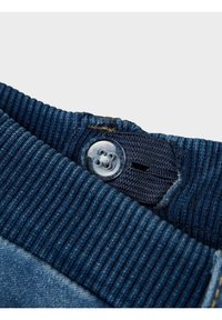 Tela de denim azul con una cinturilla de textura acanalada, que presenta un botón de plástico transparente y un ojal integrado para cerrar.