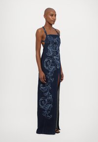 Μακριά τζιν φορεμάτα σε σκούρο μπλε χρώμα με λουλουδάτη κεντητική, πλευρικό άνοιγμα και ρυθμιζόμενες τιράντες. Λείος υφασμάτινος χαρακτήρας και κολακευτική σιλουέτα.