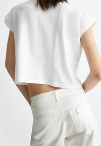 Persona que lleva una camiseta corta blanca suelta y unos pantalones vaqueros blancos de cintura alta, vista desde la espalda contra un fondo liso claro.
