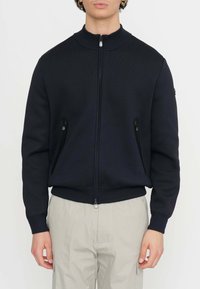 Giacca zip-up blu navy con colletto alto, polsini e orlo a coste, dotata di due tasche con cerniera e tessuto testurizzato. Gli accenti includono il logo sulla manica.