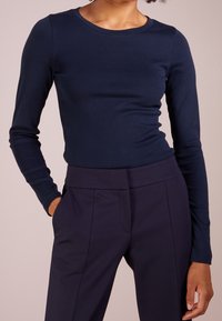 Eine Person trägt ein enges, navy-blaues Langarmshirt, das in hochtaillierte, dunkelblaue, maßgeschneiderte Hose gesteckt ist, mit den Händen in den Taschen.