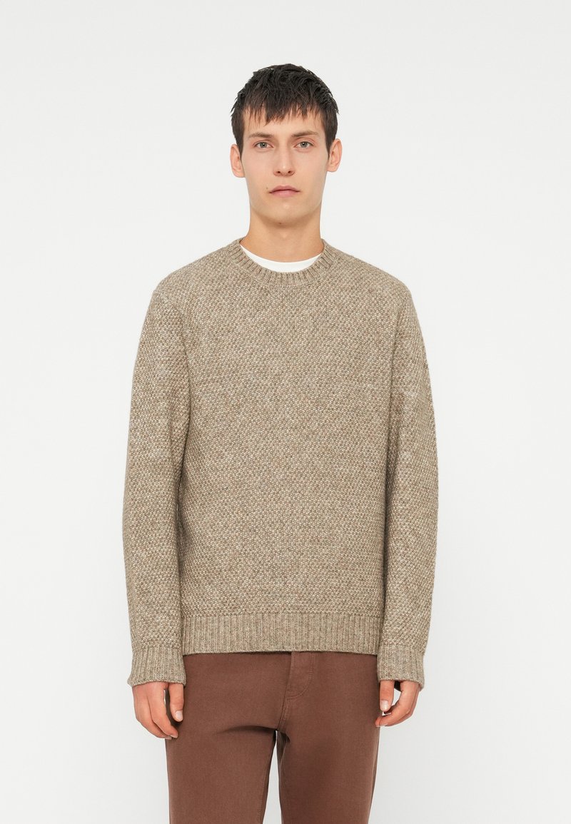 Knitted crew neck sweater in beige with a textured pattern, featuring ribbed cuffs and hem, paired with brown pants.