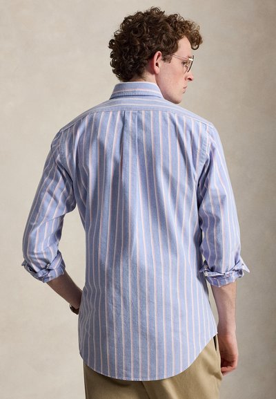 Mann iført lyseblå og hvit skjorte med vertikale striper, oppbrettede ermer og beige bukser, sett bakfra mot nøytral bakgrunn.