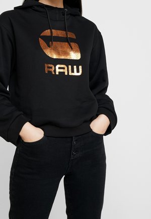Personne portant un sweat à capuche noir avec un logo "G RAW" cuivre métallique, les mains dans les poches d'un jean noir, sur un fond uni.