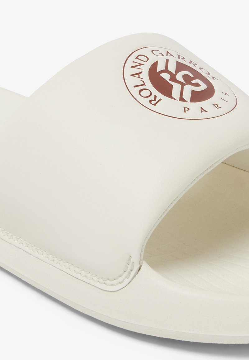 White slide sandal with a wide strap featuring a brown logo, textured footbed, and smooth synthetic material. Subtle stitching detail.