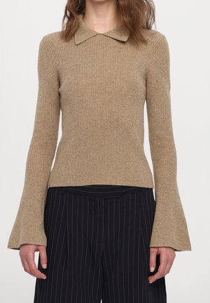 Beiger, gerippter Strickpullover mit ausgestellten Ärmeln und kleinem Kragen, kombiniert mit schwarzen Nadelstreifenhosen.