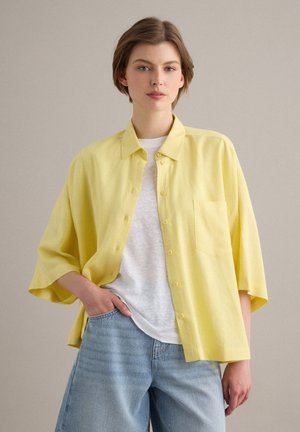 Persona joven con cabello corto y castaño lleva una camisa amarilla de gran tamaño abotonada encima de una camiseta blanca y vaqueros azul claro, de pie sobre un fondo neutro.
