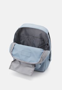 Light blue backpack with a grey flap, zip closure, and mesh side pocket. Interior has grey lining and an organizational compartment.