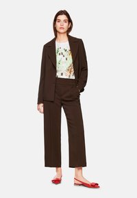 Brown tailored suit with wide-leg trousers and a fitted top featuring a geometric and botanical print. Red flat shoes with a bow accent.