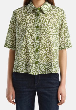 Femme portant une chemise verte à manches courtes avec des boutons et des taches blanches irrégulières, associée à un jean bleu foncé.