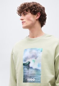 Νεαρός άνδρας με σγουρά μαλλιά που φοράει ανοιχτό πράσινο φούτερ με μεγάλο σχέδιο κυματιού της θάλασσας και τη λέξη "HUGO" μπροστά.