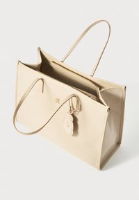 Saco tote bege feito de tecido texturizado com duas alças, um compartimento com fecho e um charme decorativo anexado.