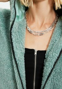 Giacca fuzzy verde menta con cerniera nera e metallizzazione argentata, sovrapposta a un top nero a coste. Collana multi-catena argentata visibile.